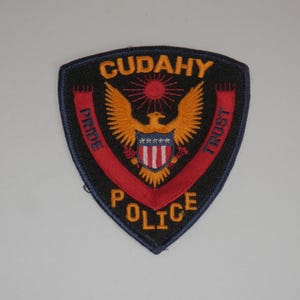 May include: A shield-shaped embroidered patch with a black background. It features a golden eagle with a red sun above, a central shield with red and white stripes and white stars, and the text CUDAHY POLICE with PRIDE and TRUST in gold.