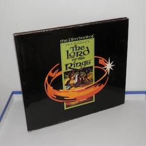 May include: A hardback book titled The Film Book of J.R.R. Tolkien's The Lord of the Rings. The black cover features a central green rectangular panel with an illustration of figures, surrounded by a stylised orange ring and a white starburst.
