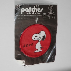 May include: A circular red self-adhesive patch featuring a white Snoopy character and the word LOVE in black text. The patch is enclosed in original black packaging with the text New! patches self-adhesive and a graphic of a person wearing a shirt.