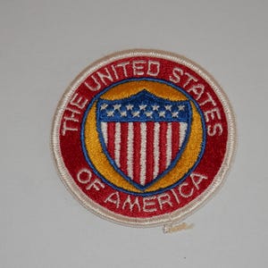 May include: A circular embroidered patch featuring a red border with the white text THE UNITED STATES OF AMERICA. Inside is a gold circle surrounding a blue shield with white stars at the top and vertical red and white stripes at the bottom.