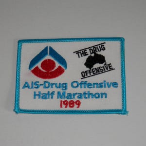 May include: A rectangular embroidered patch with a white background and a bright blue border. It features a blue and red logo, a black silhouette of Australia with the text THE DRUG OFFENSIVE, and the blue text AIS-Drug Offensive Half Marathon 1989.