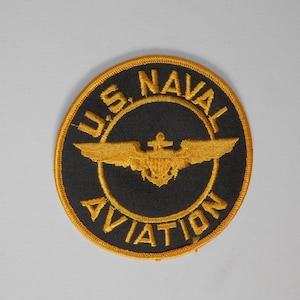 May include: A circular embroidered patch featuring a black background with a gold border. The text U.S. NAVAL AVIATION is stitched in gold thread around the centre. A gold embroidered emblem of wings and an anchor is centred in the middle.