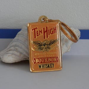 May include: A rectangular metal keyring with a tan enamel finish featuring the Ten High logo in red and gold. The design includes an eagle graphic, the text America's Native Spirit, Kentucky Straight Sour Mash Bourbon Whiskey, and a gold ring.