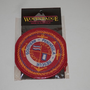 May include: A circular red embroidered patch featuring a gold rope border and two crossed anchors. The centre displays the text SOUTHERN PENINSULA RESCUE and SEMPER PARATUS with a small flag and a blue square icon on a white background.