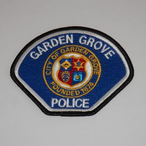 May include: A blue embroidered patch with a black border and white trim. The text GARDEN GROVE is at the top and POLICE is at the bottom. In the centre is a circular emblem with the text CITY OF GARDEN GROVE FOUNDED 1874 and a shield.