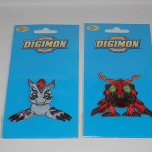 May include: Two Digimon embroidered patches on blue backing cards. The left patch features a grey creature with a red crest and green eyes. The right patch shows a red, beetle-like creature with green eyes and orange antennae. Both cards feature the Digimon logo.