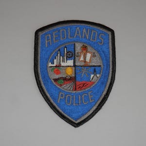 May include: A shield-shaped embroidered patch with a blue background and black border. The text REDLANDS POLICE is stitched in grey. A central circular emblem features four quadrants depicting a city skyline, a scale of justice, fruit, and a star.