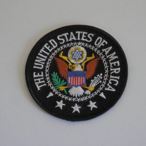 May include: A circular embroidered patch with a black background featuring the Seal of the President of the United States. The text THE UNITED STATES OF AMERICA is stitched in white around the top, with three white stars at the bottom.