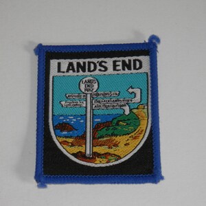 May include: A rectangular embroidered patch with a blue border featuring a graphic of a signpost at Land's End. The design includes a light blue sky, a dark blue sea, and a sandy beach. The text reads LAND'S END at the top and on the signpost.