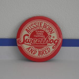 May include: A circular red and cream pinback button featuring the text "AUSSIE BORN AND BRED" in a circular border, with "TOUGH TIMES CLOTHES" and "Sweathog" printed in the centre in a stylised red font against a cream background.