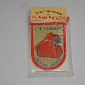 May include: A rectangular woven badge in a clear plastic sleeve with a header card reading Barker Souvenirs WOVEN BADGES. The badge features a tan background, a red border, and a graphic of a red mountain with the text I'VE CLIMBED AYERS ROCK.