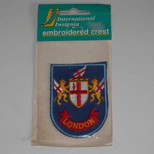 May include: An embroidered crest patch featuring the London coat of arms on a blue shield, flanked by two gold lions holding flags with red crosses. A red banner at the bottom displays the text LONDON. The patch is in a clear plastic retail package.