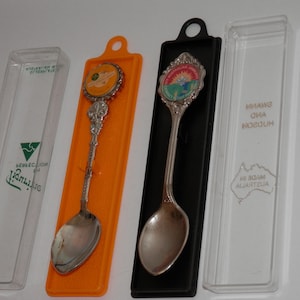 May include: Two silver-toned souvenir spoons with decorative handles and circular emblems at the top. One spoon rests in an orange plastic holder, the other in a black plastic holder. Two clear plastic cases are included, one marked with the text Figurine.
