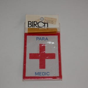 May include: A rectangular embroidered patch in a clear plastic package with a Birch brand header card. The patch features a white background, a red border, a central red cross, and the blue text PARA above and MEDIC below the cross.