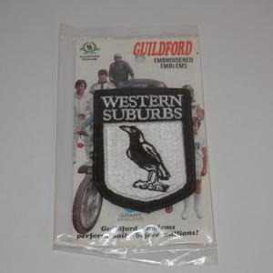 May include: A shield-shaped embroidered patch featuring a black magpie on a white background with the text WESTERN SUBURBS in white on a black border. The patch is inside a clear plastic package with a backing card labeled GUILDFORD EMBROIDERED EMBLEMS.