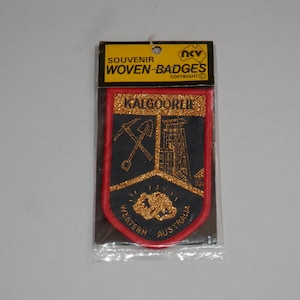 May include: A souvenir woven badge in a plastic package with a header card reading SOUVENIR WOVEN-BADGES. The shield-shaped patch features a red border, a black background, and gold embroidery depicting a pickaxe, shovel, and mining structure.