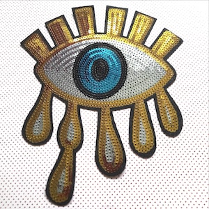 Op de afbeelding: Een gouden en zilveren paillettenoog met een blauwe pupil en gouden traanvormen die eronder hangen.