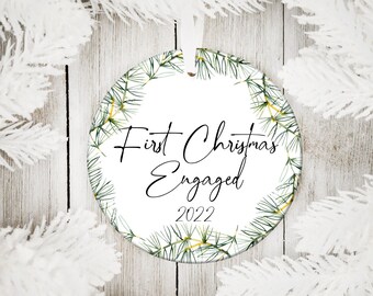 Puede incluir: Adorno navideño redondo blanco con un diseño de rama de pino verde y el texto "First Christmas Engaged 2022".