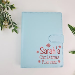 Puede incluir: Una agenda azul claro con un diseño de copo de nieve rojo y el texto "Sarah's Christmas Planner" en la portada.