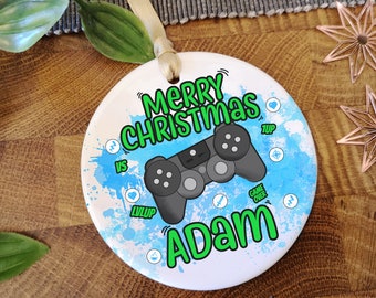 May include: White round Christmas ornament with a blue and green watercolor background. The ornament features a black video game controller and the text "Merry Christmas Adam" in green.