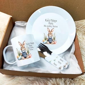 Könnte beinhalten: Eine weiße Keramiktasse und ein Teller mit einem Hasendesign und dem Text "Kalo Pascha Aris Me agapi, Nouna". Tasse und Teller befinden sich in einem braunen Karton mit weißem Seidenpapier. Außerdem sind zwei weiße Plastikgabeln mit Hasendesign und dem Text "Aris" auf den Griffen enthalten.