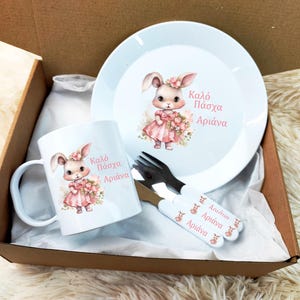Könnte beinhalten: Ein weißes Oster-Geschenkset mit einem Hasendesign. Das Set enthält eine Tasse, einen Teller und eine Gabel. Tasse und Teller zeigen einen rosa Hasen mit dem Text "Kaló Páxcha Ariádna". Die Gabel hat ein rosa Hasendesign und den Text "Ariádna" dreimal wiederholt.