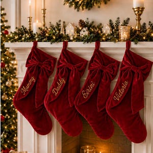 以下が含まれることがあります： 大きな装飾リボンが付いた、深紅のベルベット製クリスマスストッキング4足が並んで吊るされています。各ストッキングには、Mummy、Daddy、Eirini、Victoriaという名前が金色の筆記体で刺繍されています。