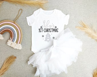 May include: White baby onesie with a silver glitter design that says "Lily's 1st Christmas" with a small silver glitter Christmas tree. The onesie is paired with a white tulle tutu.