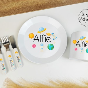 May include: A white children's dinnerware set with a space theme. The set includes a plate, bowl, cup, fork, knife, and spoon. The plate, bowl, and cup feature a colorful design of planets, stars, and a rocket ship. The name "Alfie" is printed on each piece. The set is made of durable polymer.