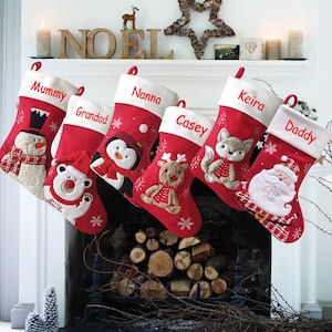 Op de afbeelding: Zes rode kerstsokken met witte bies en geborduurde namen: Mummy, Grandad, Nanna, Casey, Keira en Daddy. Elke sok heeft een andere geborduurde applicatie van een sneeuwpop, ijsbeer, pinguïn, rendier, vos en kerstman.