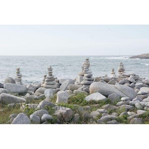 Peut inclure: Paysage côtier avec plusieurs empilements de pierres grises, ressemblant à de petites tours, sur une plage rocheuse. L'océan est visible en arrière-plan sous un ciel pâle. De la végétation verte est dispersée parmi les rochers.