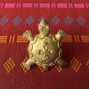 Könnte beinhalten: Eine kleine, goldfarbene Schildkrötenfigur mit einem detaillierten Design auf ihrem Panzer. Die Schildkröte sitzt auf einem roten und orangefarbenen gemusterten Stoff.
