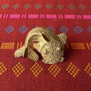 Könnte beinhalten: Eine goldfarbene Metallfischfigur mit einer detaillierten, strukturierten Oberfläche. Der Fisch liegt auf einem roten und orangefarbenen gemusterten Stoff.