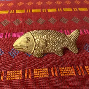 Könnte beinhalten: Ein goldfarbenes, fischförmiges Dekorationsobjekt mit einer strukturierten Oberfläche. Der Fisch liegt auf einem roten Stoff mit geometrischem Muster.