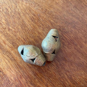 May include: Two gold-coloured metal beads with a circular pattern. The beads are on a wooden surface.