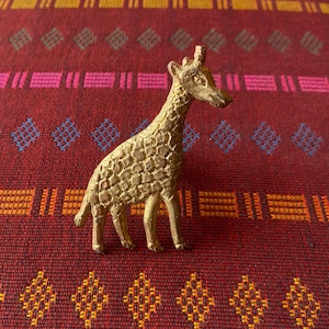 以下が含まれることがあります： テクスチャ加工された表面を持つ金色のキリンの置物。置物は赤とオレンジのパターンの布の上に置かれています。