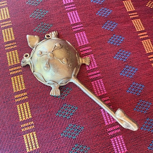 Könnte beinhalten: Ein dekorativer, goldfarbener Metalllöffel mit einer verzierten, runden Schüssel und einem langen Griff. Der Löffel hat ein detailliertes Design und liegt auf einem gemusterten roten und kastanienbraunen Stoff.