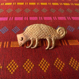 Peut inclure: Une broche en métal de couleur dorée en forme de caméléon stylisé. La broche a une surface texturée et une queue enroulée.
