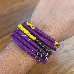 Könnte beinhalten: Ein Set aus sechs violetten Stoffarmbändern mit gelben und grünen Mustern. Die Armbänder sind auf dem Handgelenk einer Person gestapelt.