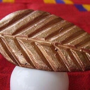 Op de afbeelding: Een bronzen, bladvormig decoratief object. Het blad heeft gedetailleerde nerven en rust op een witte, afgeronde basis. De achtergrond is een rode stof met gele en blauwe accenten.