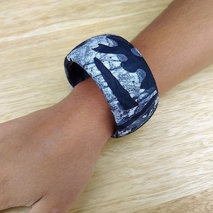 Könnte beinhalten: Ein schwarzes und weißes Armband mit Muster, mit einem breiten, flachen Design. Das Armband ist aus einem glatten, harten Material gefertigt und hat eine leicht strukturierte Oberfläche.