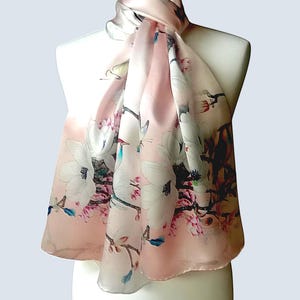 Peut inclure: Une écharpe en soie rose clair avec un motif floral de fleurs blanches et roses. L'écharpe est drapée sur un mannequin blanc, mettant en valeur son design délicat et sa palette de couleurs douces. Les dimensions de l'écharpe ne sont pas visibles sur l'image.