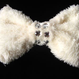 Op de afbeelding: Een witte, pluche strik met een cluster van heldere strass steentjes in het midden.