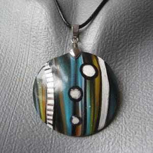 Peut inclure: Un collier avec un pendentif rond et un cordon en cuir noir. Le pendentif est en pâte polymère et présente un motif abstrait coloré avec des rayures blanches, noires, jaunes, orange, bleues et turquoise. Il y a trois cercles blancs sur le pendentif.