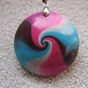 Può includere: Un ciondolo rotondo con un design a spirale nei toni del rosa, blu, marrone e grigio. Il ciondolo è attaccato a un cordino nero con una chiusura in metallo color argento. Il ciondolo è su una superficie bianca strutturata.