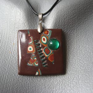 May include: A brown and green square pendant necklace with a geometric pattern and a green glass bead. The pendant is attached to a black cord.