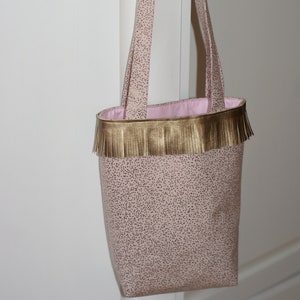 May include: A pink tote bag with gold fringe and a pink polka dot pattern. The bag has two long straps.