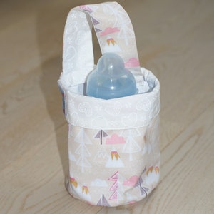 May include: A beige fabric baby bottle holder with a white trim and a pink and gray mountain and cloud pattern. The holder has a white handle and a clear baby bottle inside.