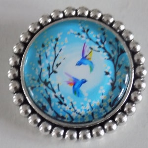Runde Silberbrosche, Glascabochon mit Vogel- und Blumenmotiv, Farben Blau/Weiß/Schwarz.