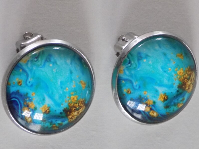 May include: A pair of silver clip-on earrings with a round blue, white, and gold abstract design.